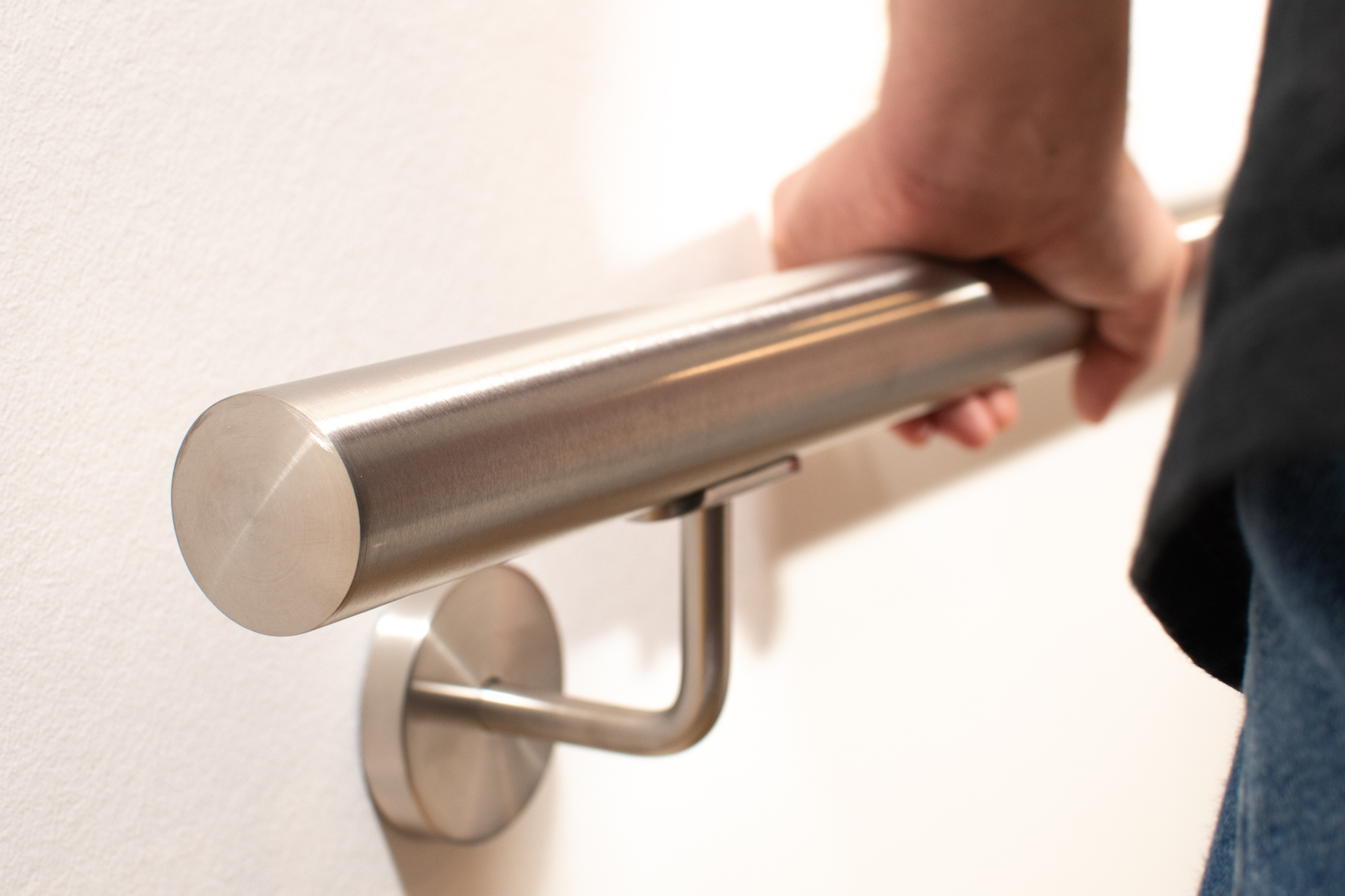 Roestvrijstalen leuning, rond, aan witte wand met roestvrijstalen houder, hand pakt de leuning, vlakke kopse kant vooraan
