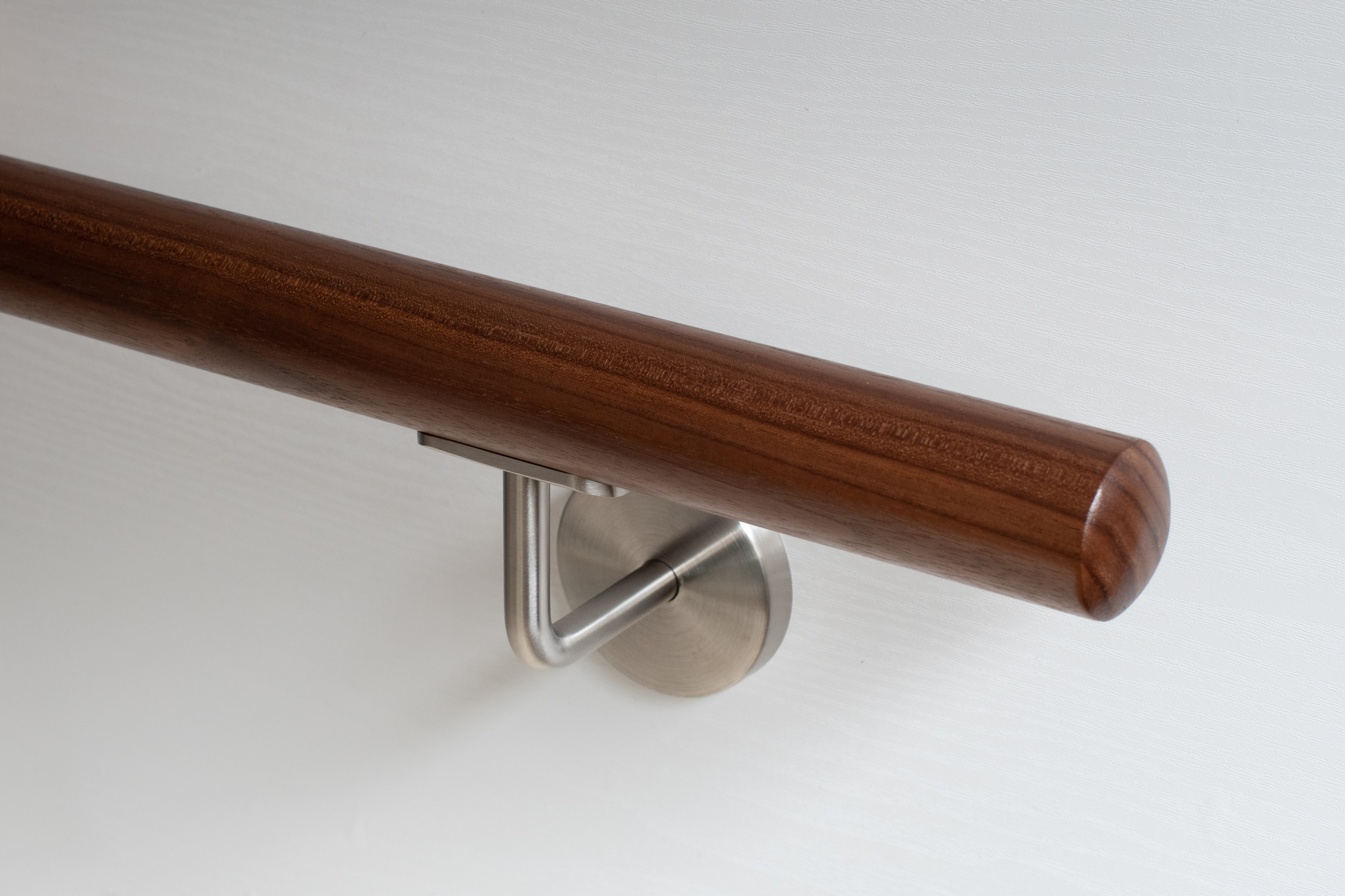 Ronde leuning van notenhout met roestvrijstalen wandhouder aan witte wand, donkerbruin hout, close-up van het uiteinde