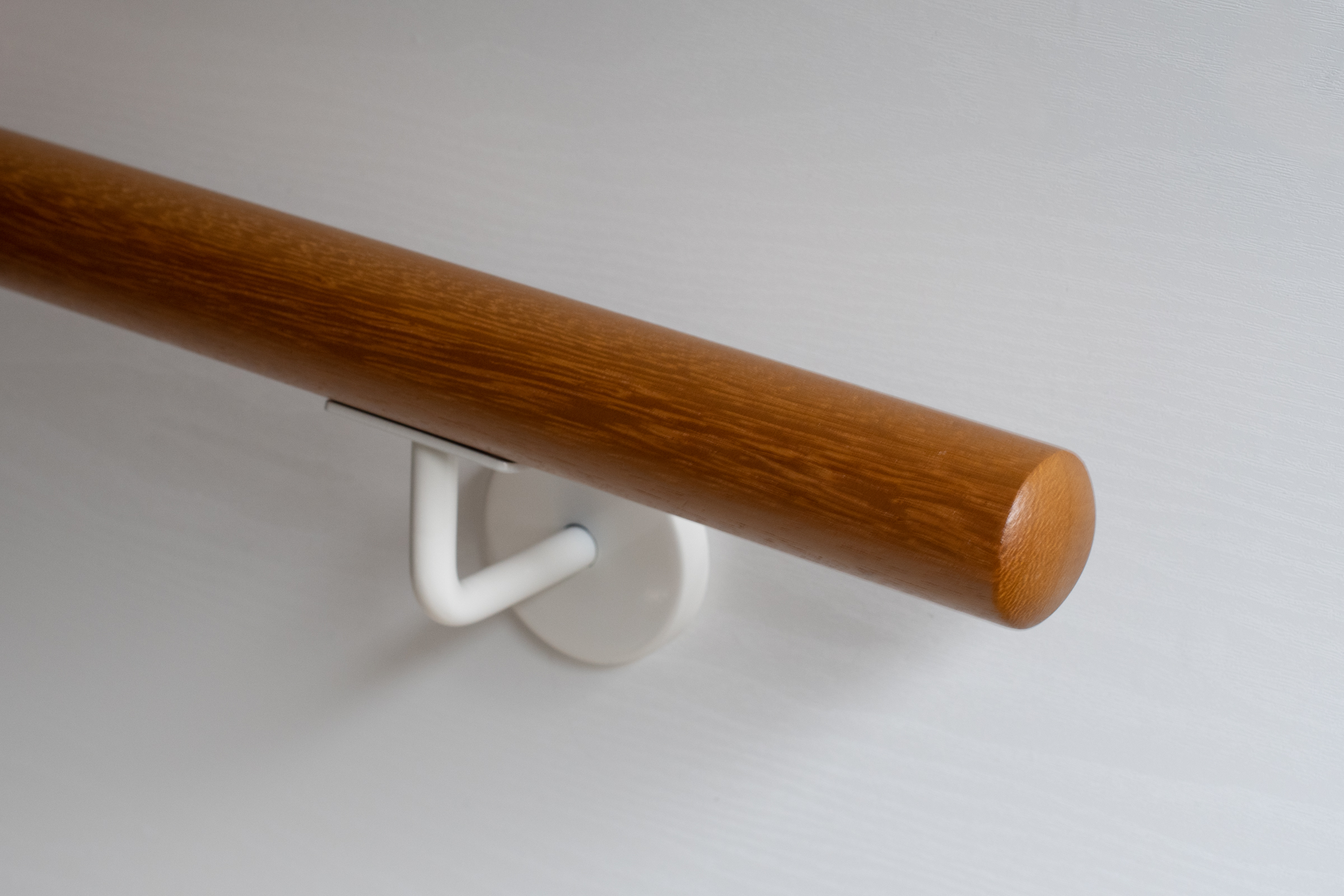 Ronde leuning van iroko met wit gelakte wandhouder aan witte wand, goudbruin hout, close-up van het uiteinde