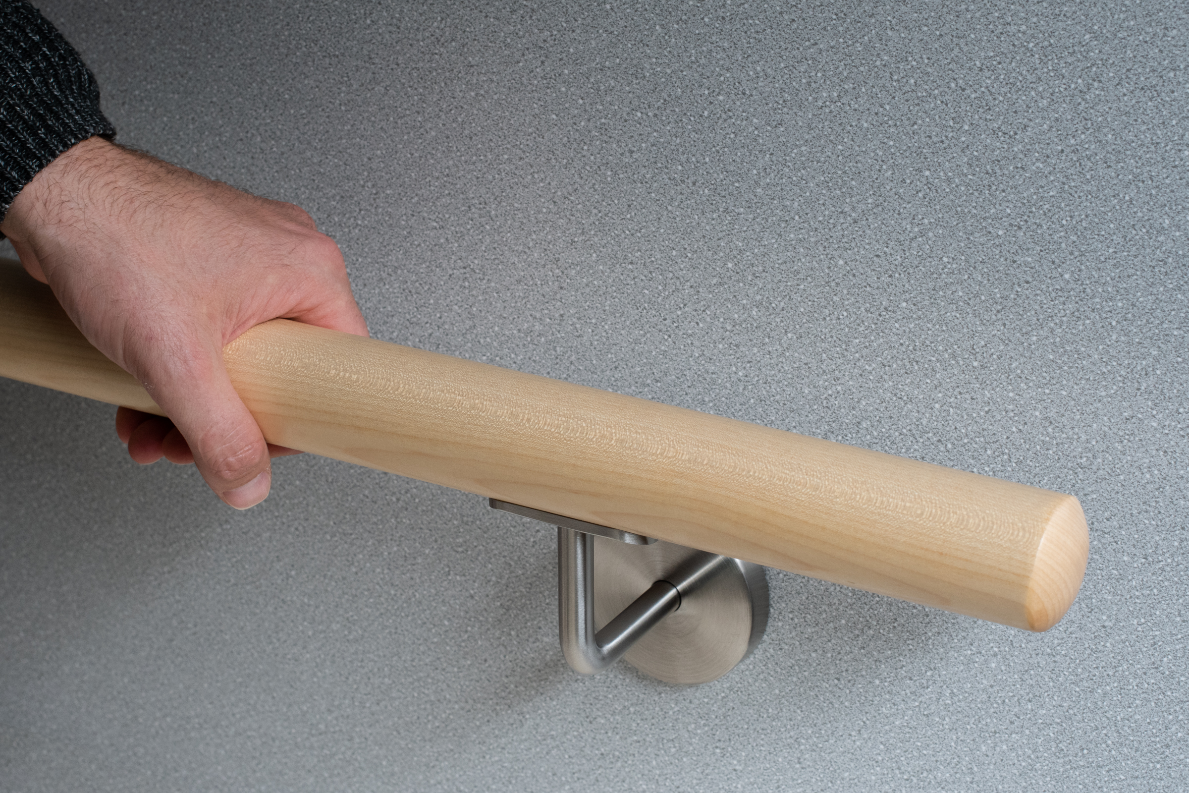 Ronde leuning van esdoornhout met roestvrijstalen wandhouder voor grijze wand, hand omvat de lichte houten leuning, close-up