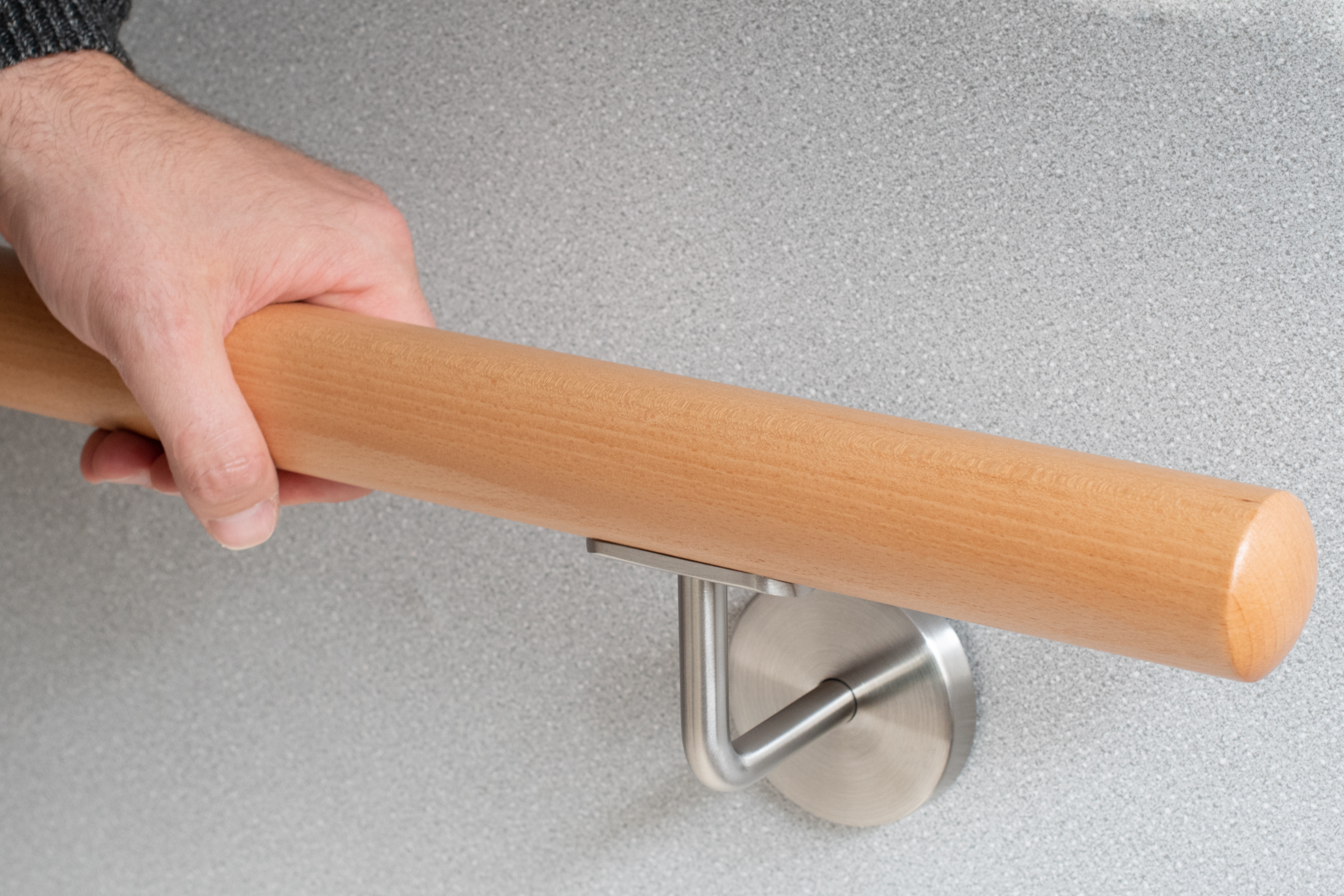Ronde leuning van beukenhout aan grijze wand met roestvrijstalen leuninghouder, hand pakt de leuning vast, gripdetail