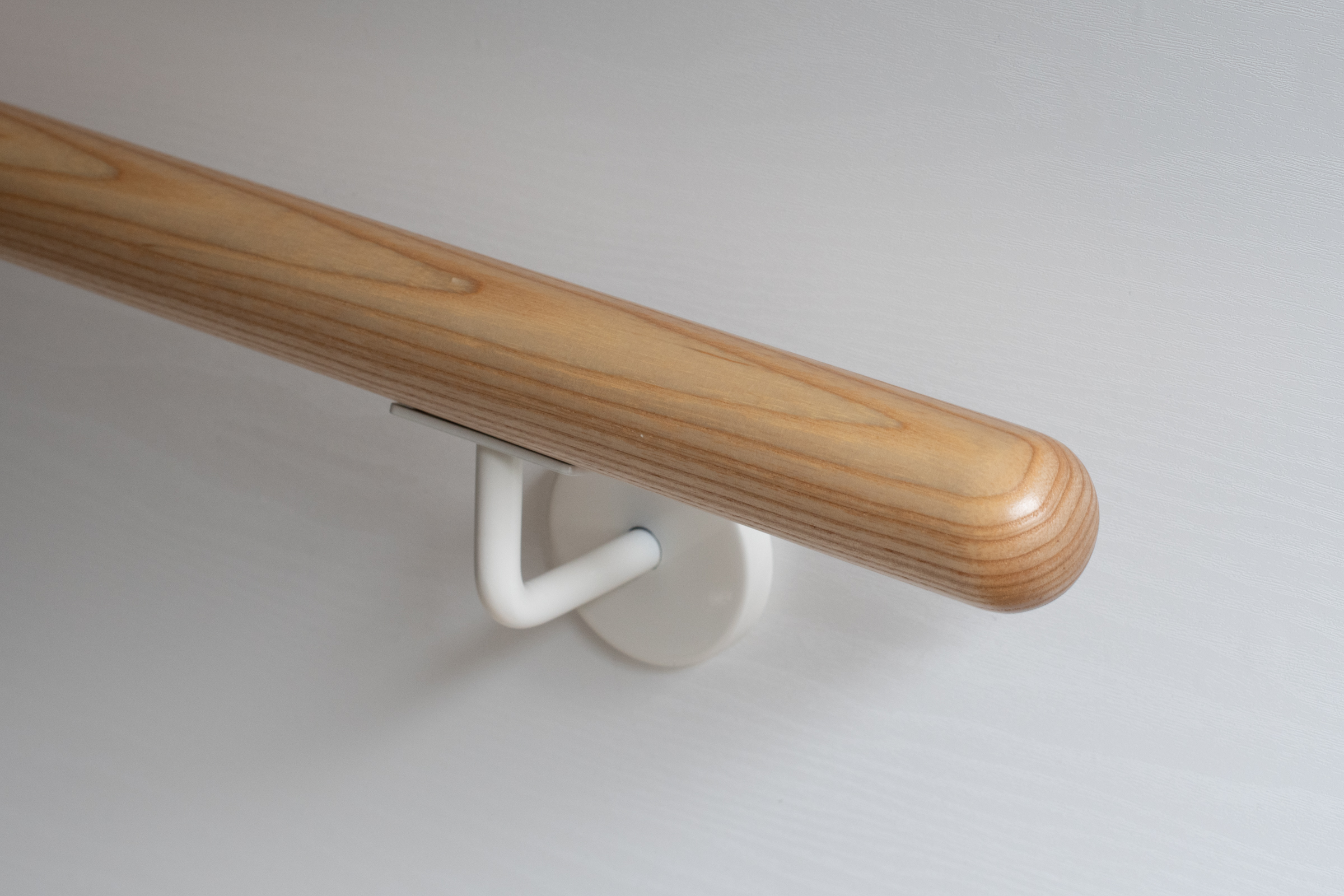 Ronde leuning van essenhout met wit gelakte wandhouder aan witte wand, licht hout met opvallende nerf, close-up