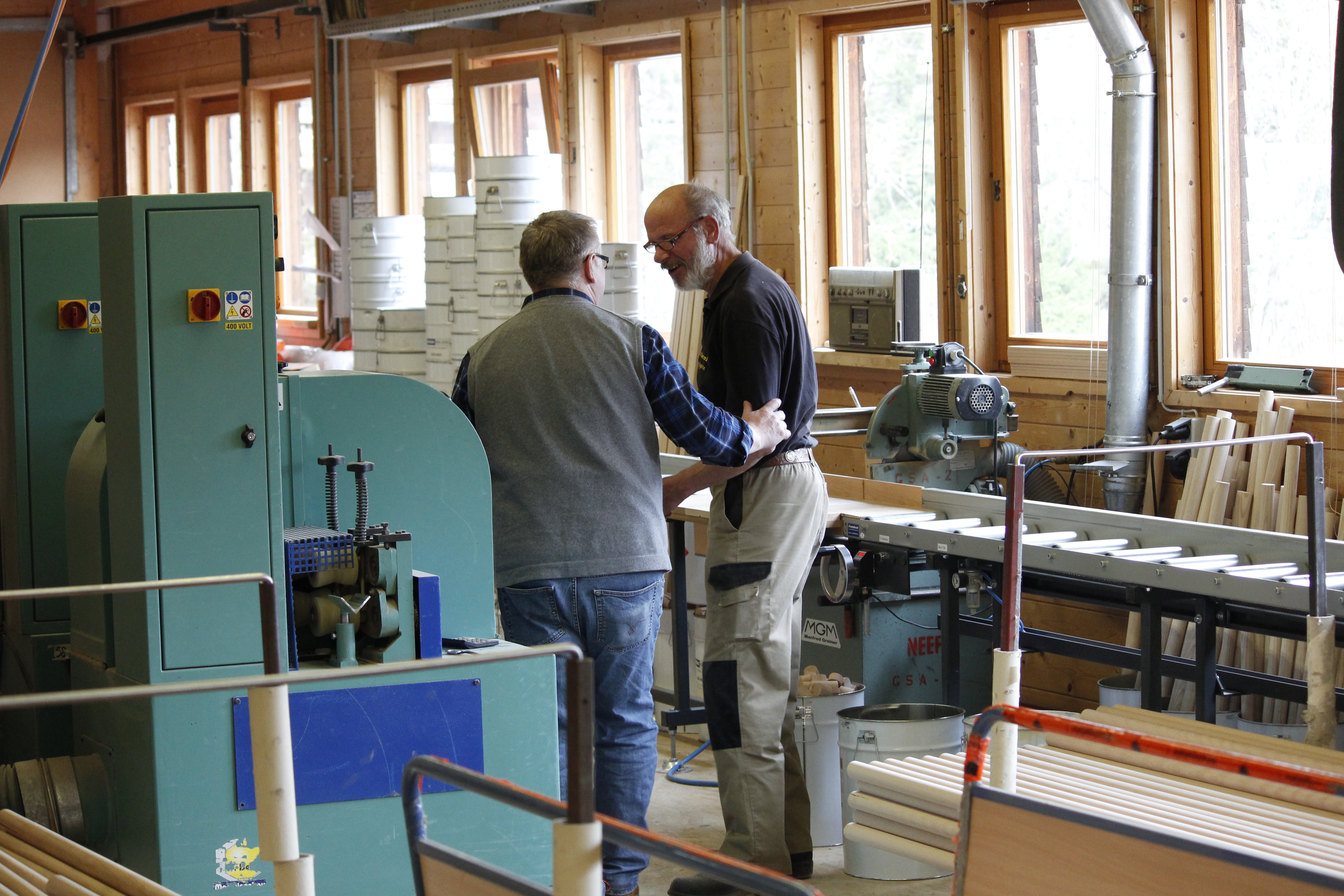 Twee medewerkers van de houtwerkplaats in gesprek tussen machines, raampartij en houtmateriaal op de achtergrond
