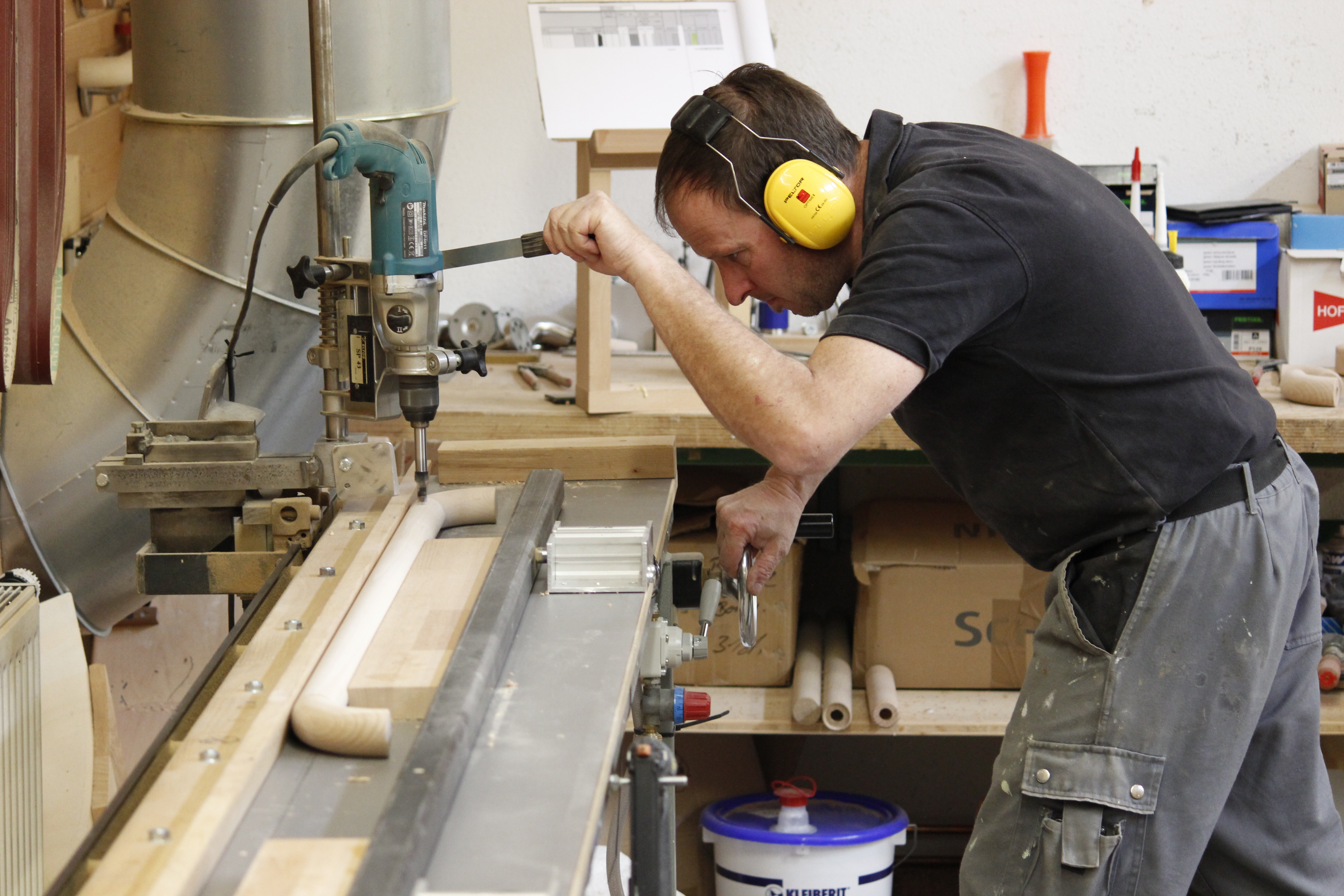 Vakman met gehoorbescherming boort in de werkplaats met een kolomboormachine gaten in een houten leuning