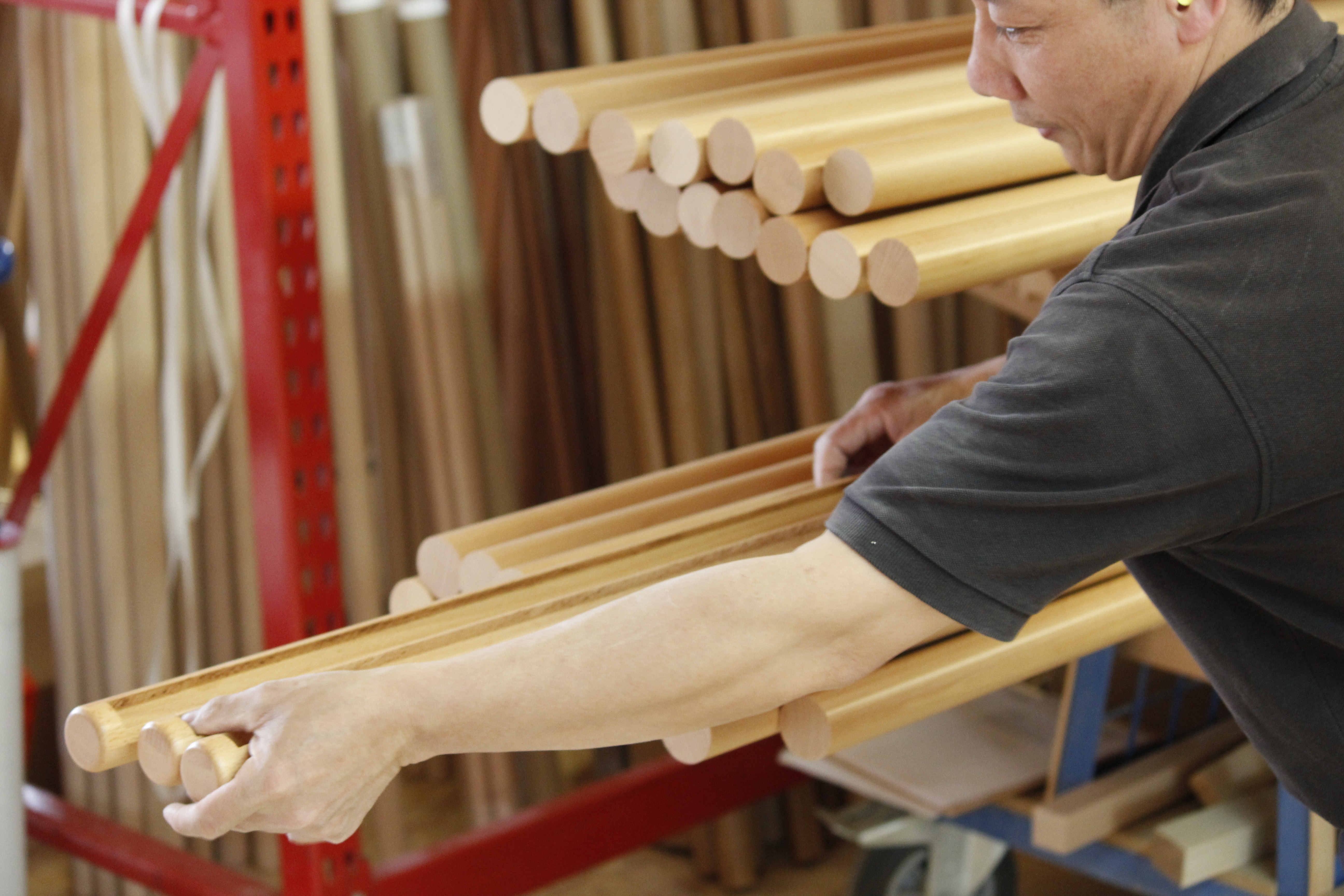 Medewerker draagt een bundel ronde houten leuningen van beukenhout door de werkplaats, rek met houten staven erachter