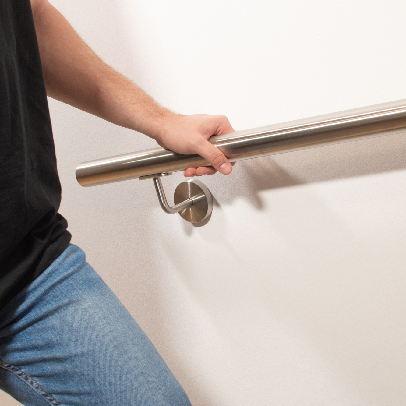 Ronde roestvrijstalen leuning aan witte wand met roestvrijstalen leuninghouder, persoon in jeans houdt zich vast aan leuning