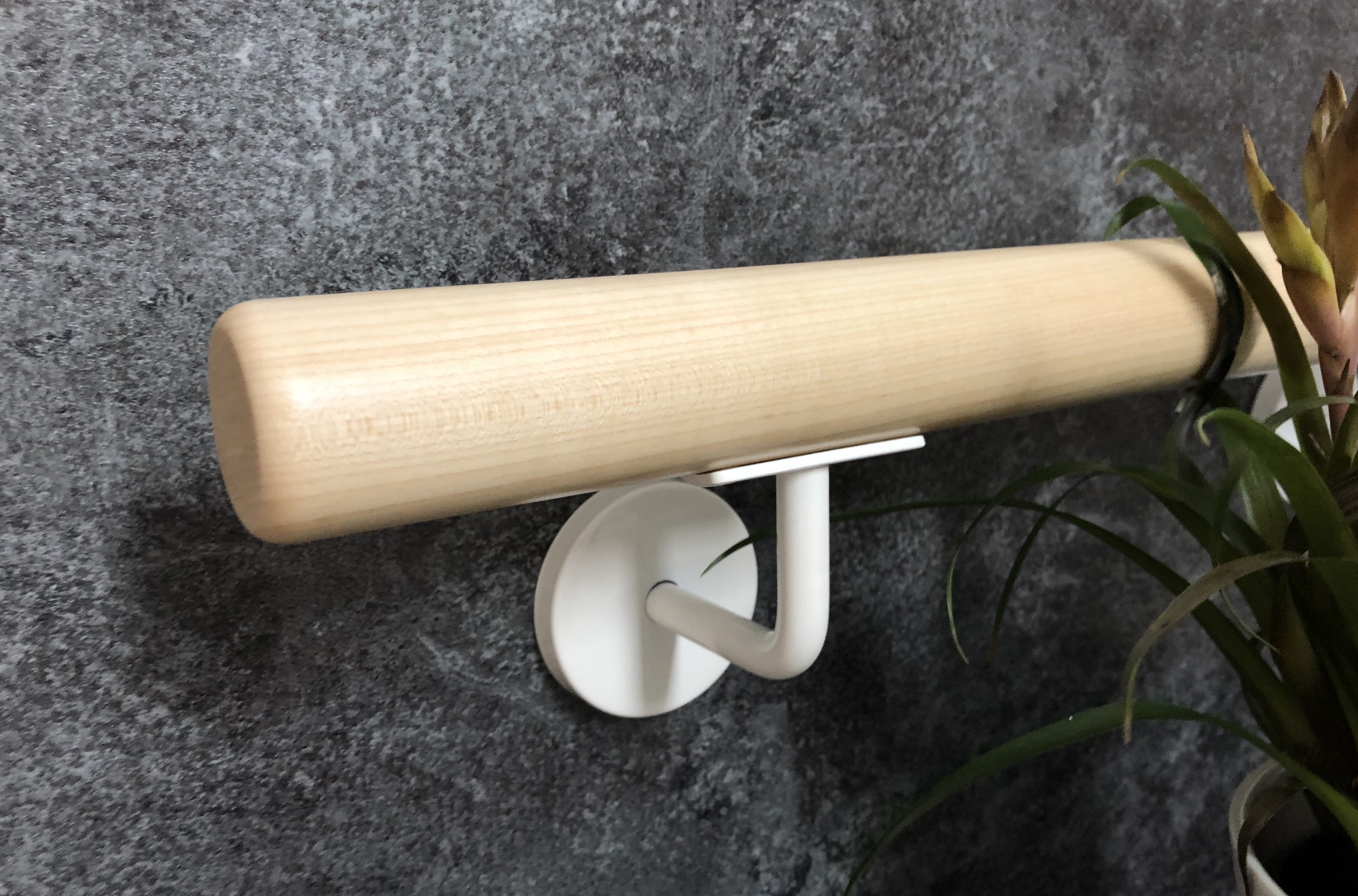 Ronde houten leuning van licht hout met wit gelakte leuninghouder aan donkergrijze stenen wand, groene plant aan de beeldrand