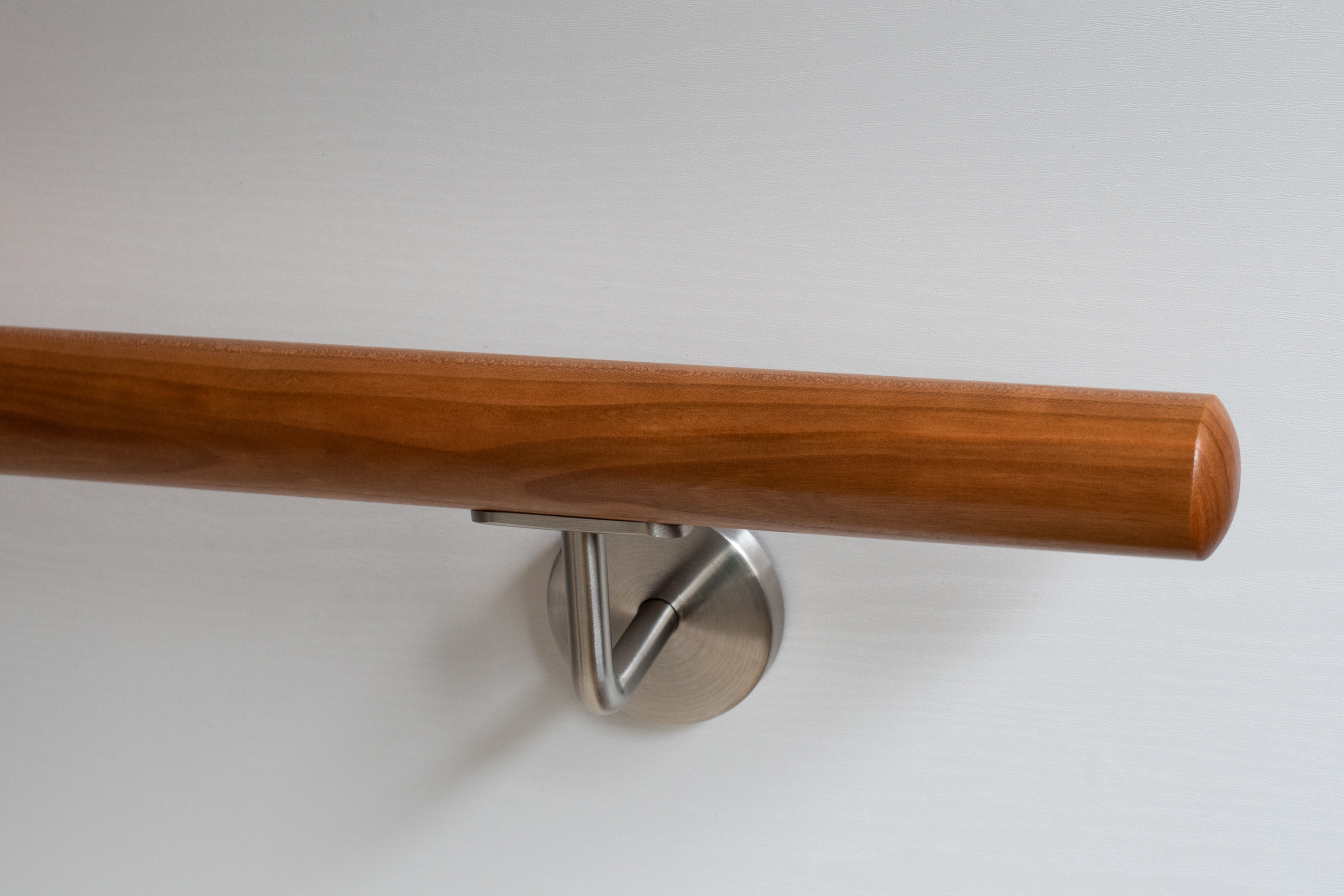 Ronde leuning van kersenhout met roestvrijstalen wandhouder aan witte wand, warm roodbruin hout, close-up van het uiteinde